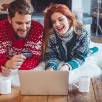 Couple on vacation at mountain cabin. Sitting on the floor on a blanket by a fireplace in a cozy living room on Christmas. Using laptop to buy gifts for friend and family. Wearing festive knitted sweaters. Austrian Alps.